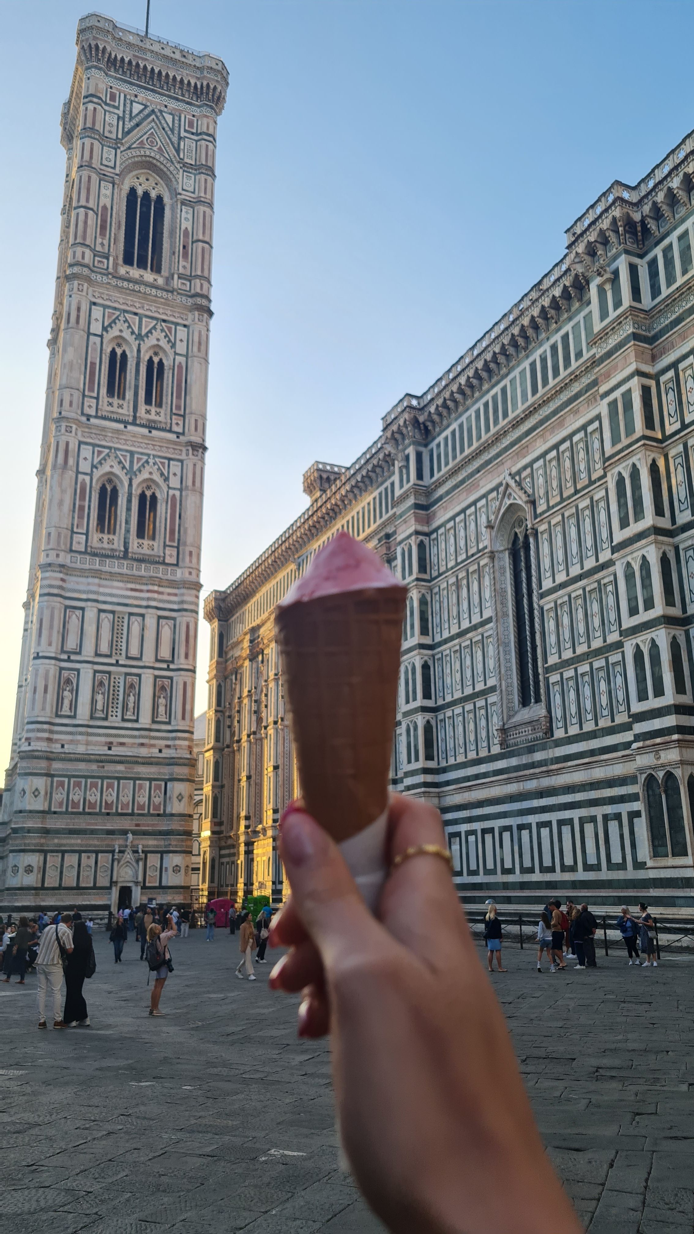 A hand holding a pink gelato cone in front of Giotto's Bell Tower and the Duomo at sunset, Florence