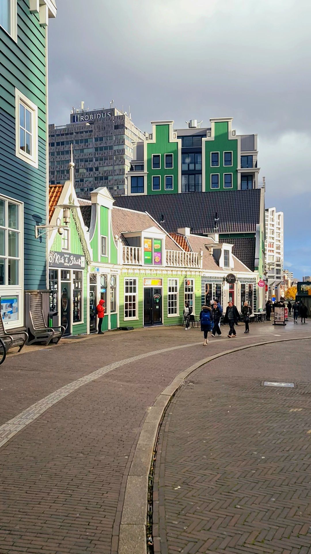 Green and white gabled shopfronts along a curved brick street with a modern tower behind them, Zaandam