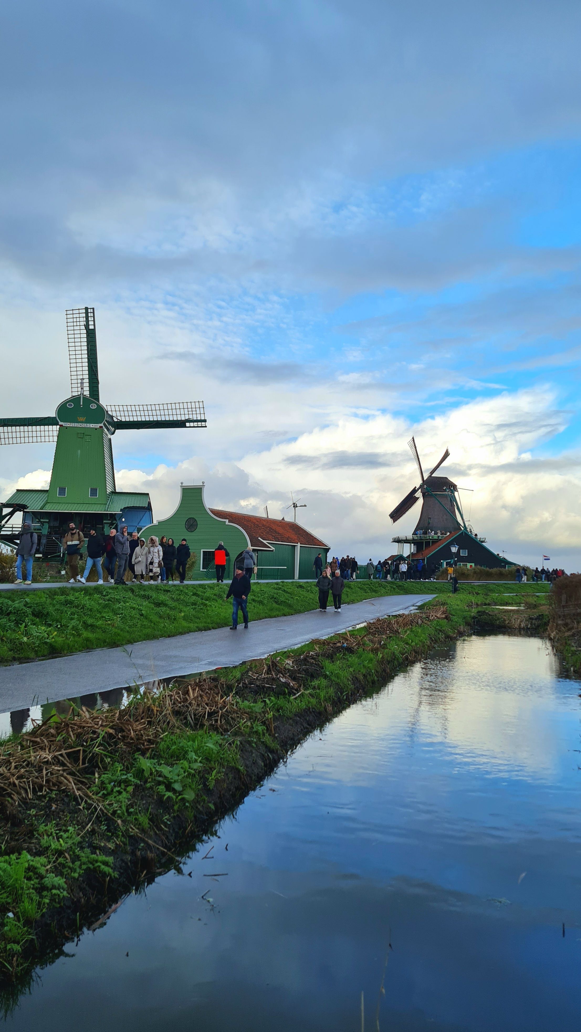 A green windmill and a dark windmill on either side of a path and a reflective ditch under blue sky and clouds, Zaanse Schans