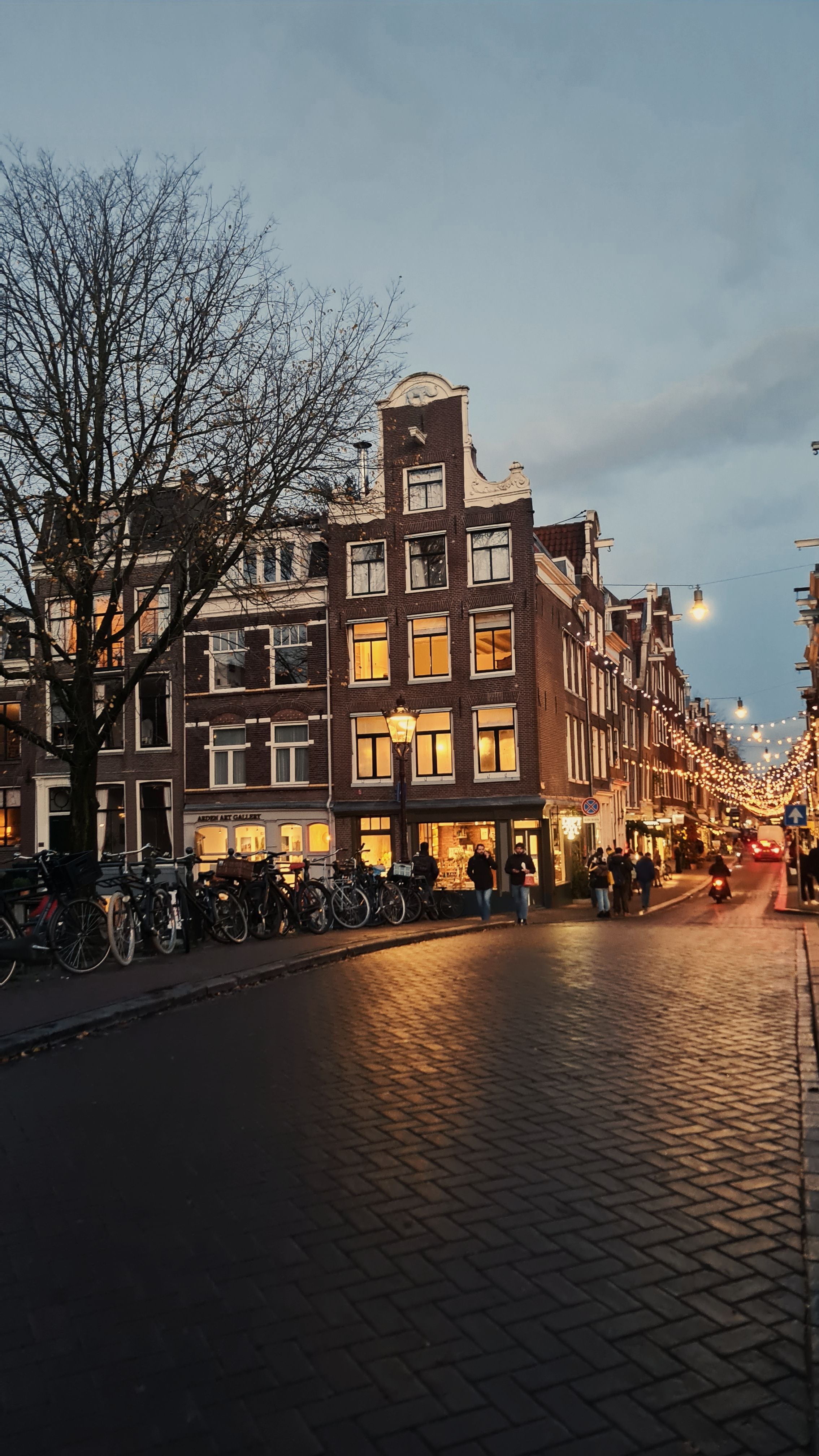 A brick corner house with glowing windows at dusk beside a street strung with fairy lights, Amsterdam
