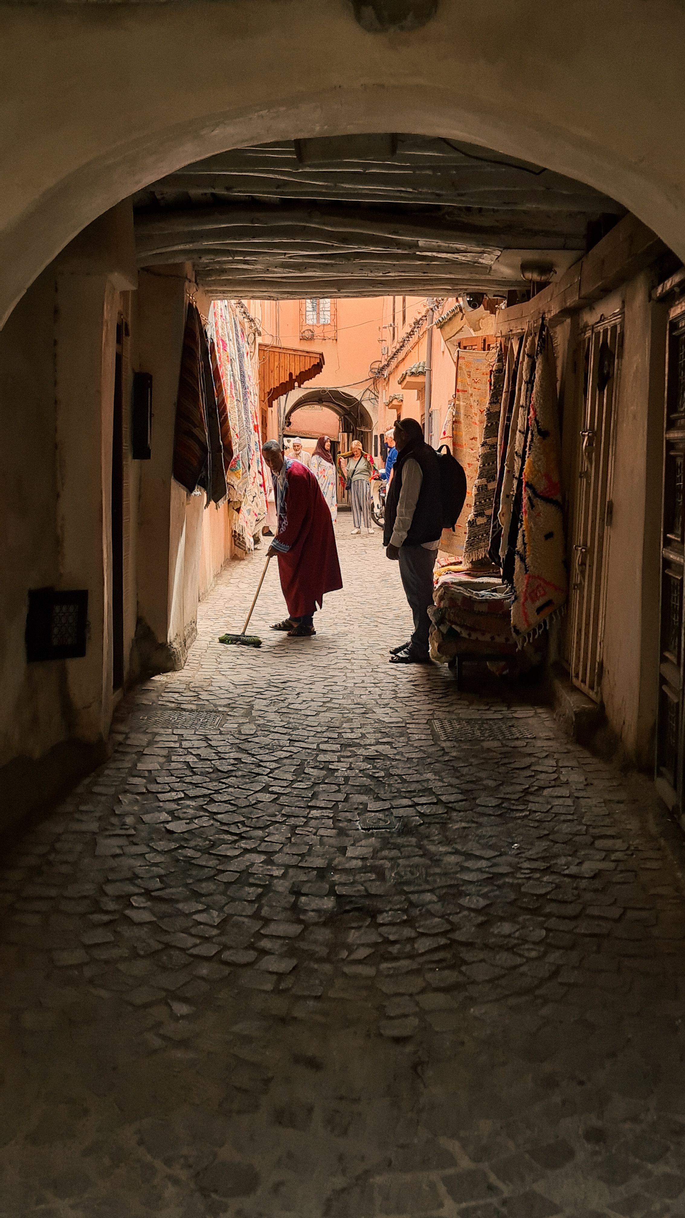 A man sweeping a cobbled medina alley under a stone arch, with rugs hanging on the walls, Marrakech