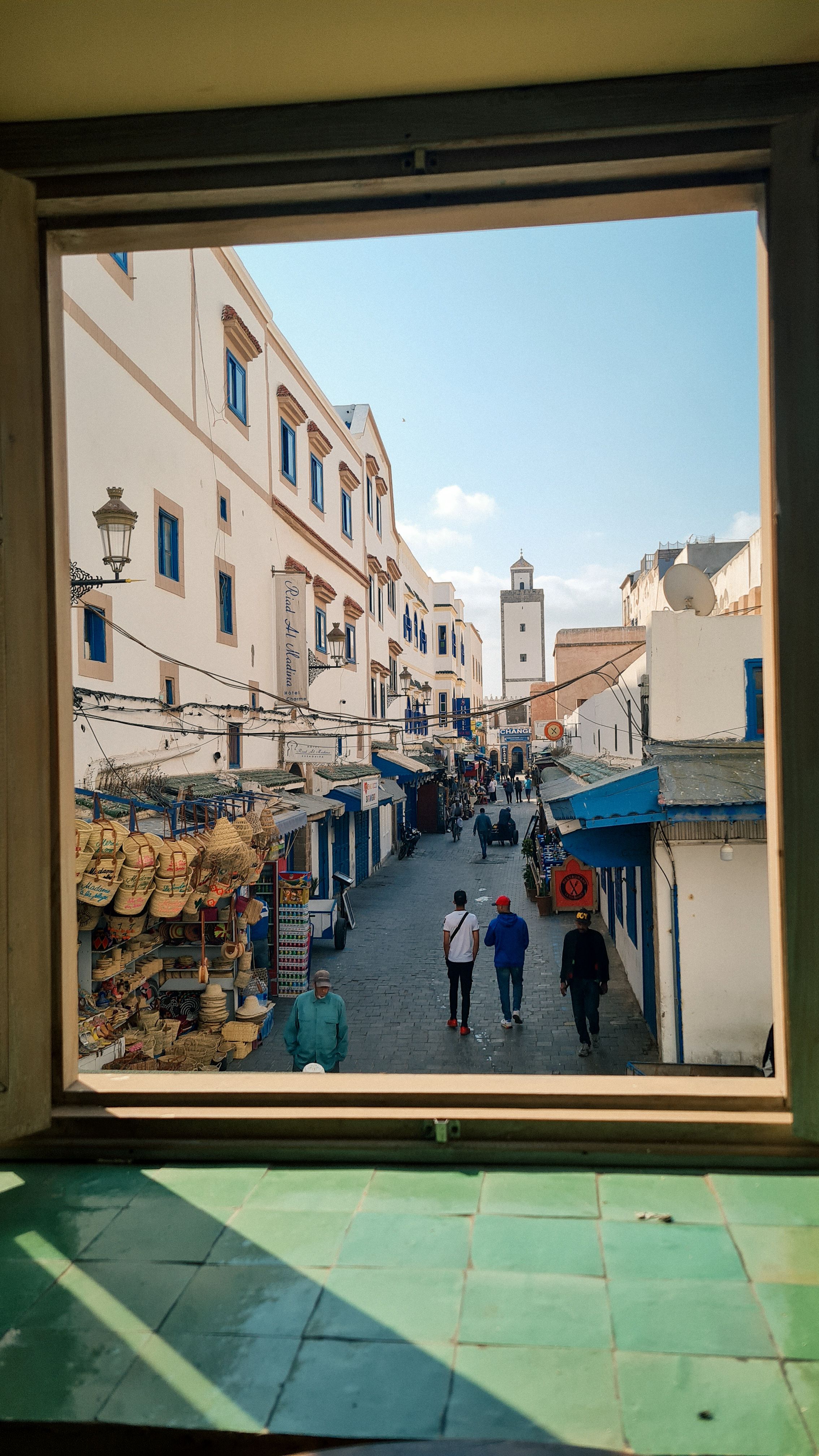 A whitewashed Essaouira street with straw hats and a minaret, framed by an open window, Essaouira