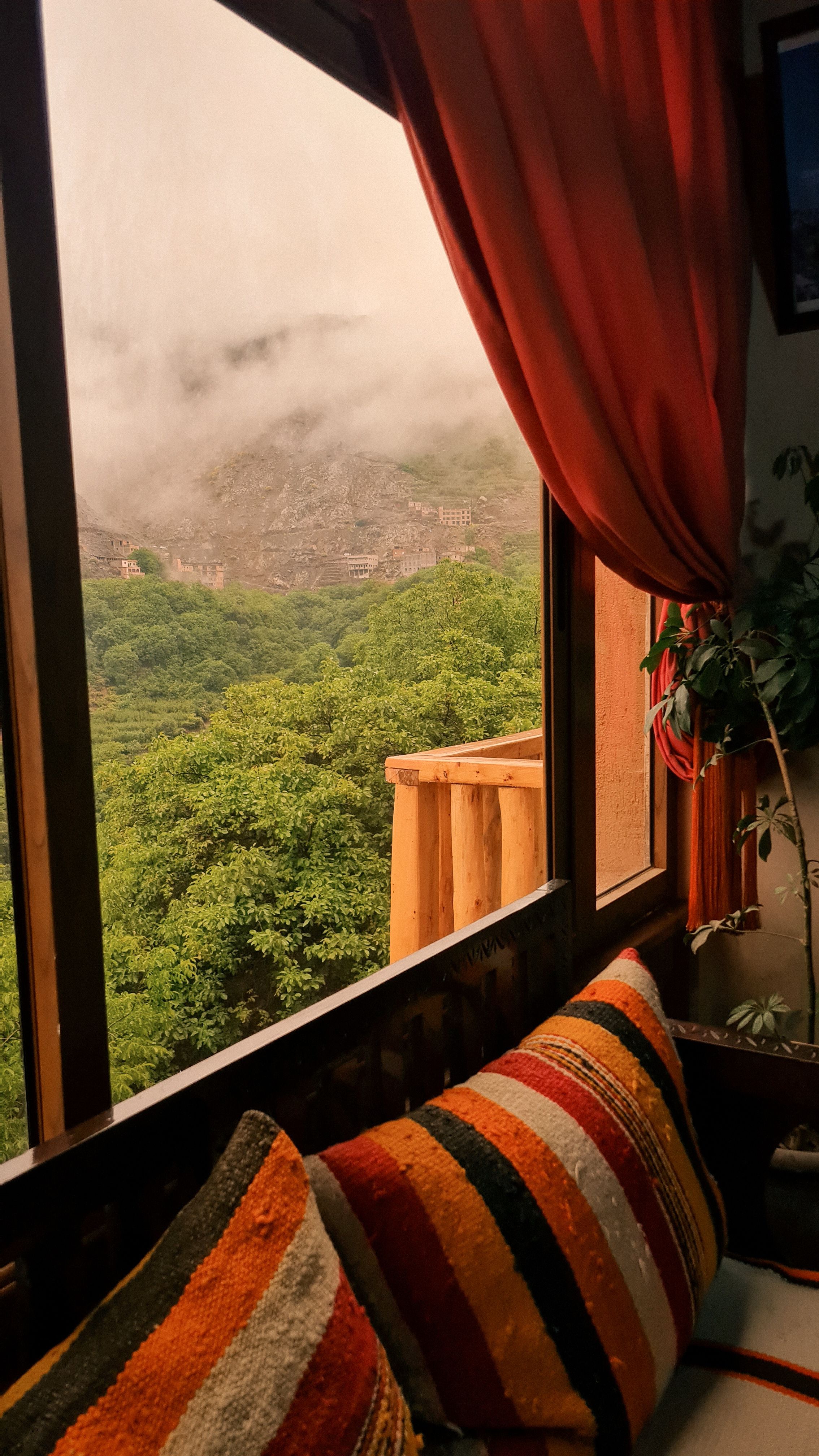 A window with orange curtains and striped cushions looking out on a misty green mountainside, Imlil