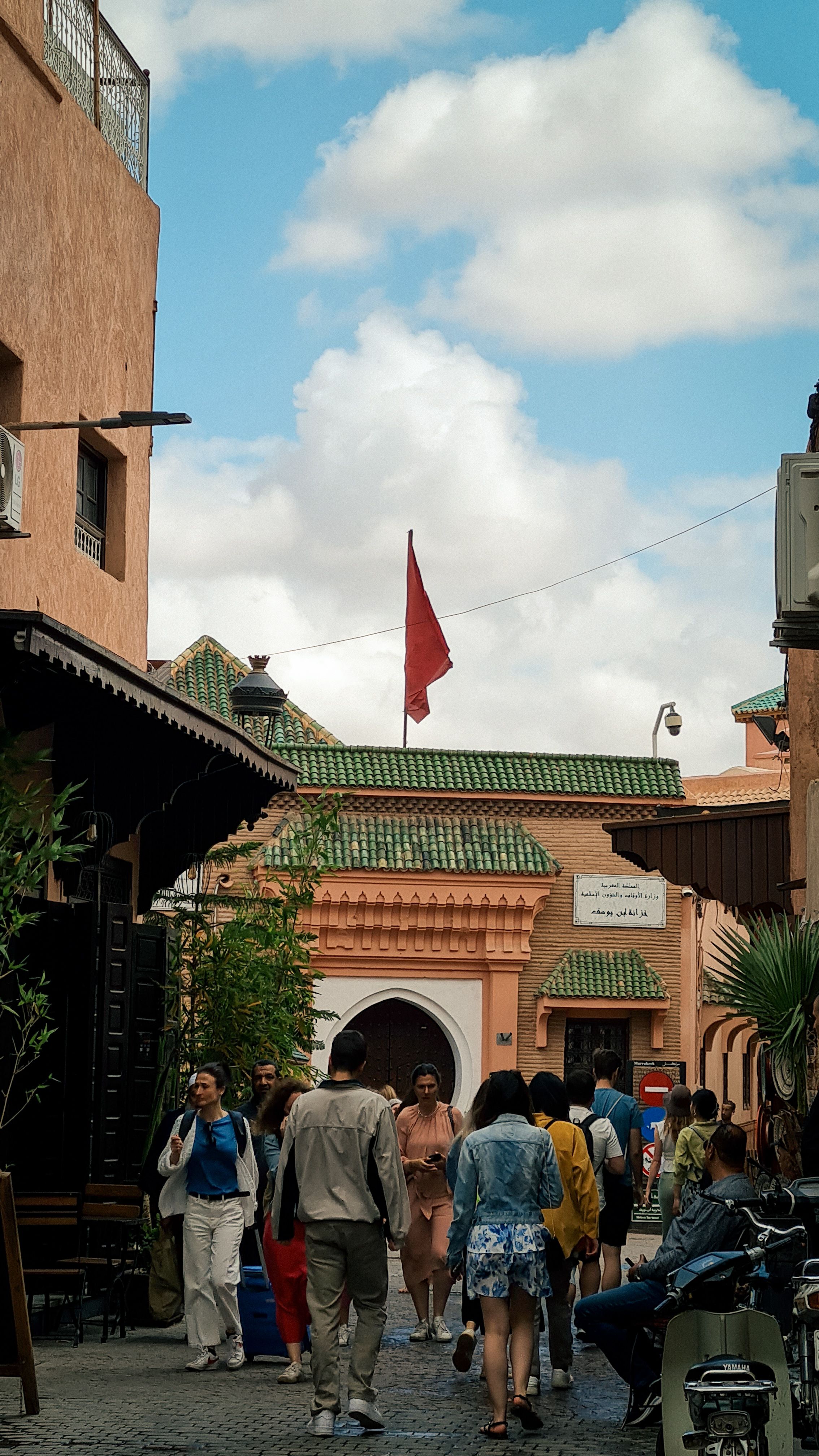 A crowded medina street below a green-tiled roof and a red Moroccan flag, Marrakech