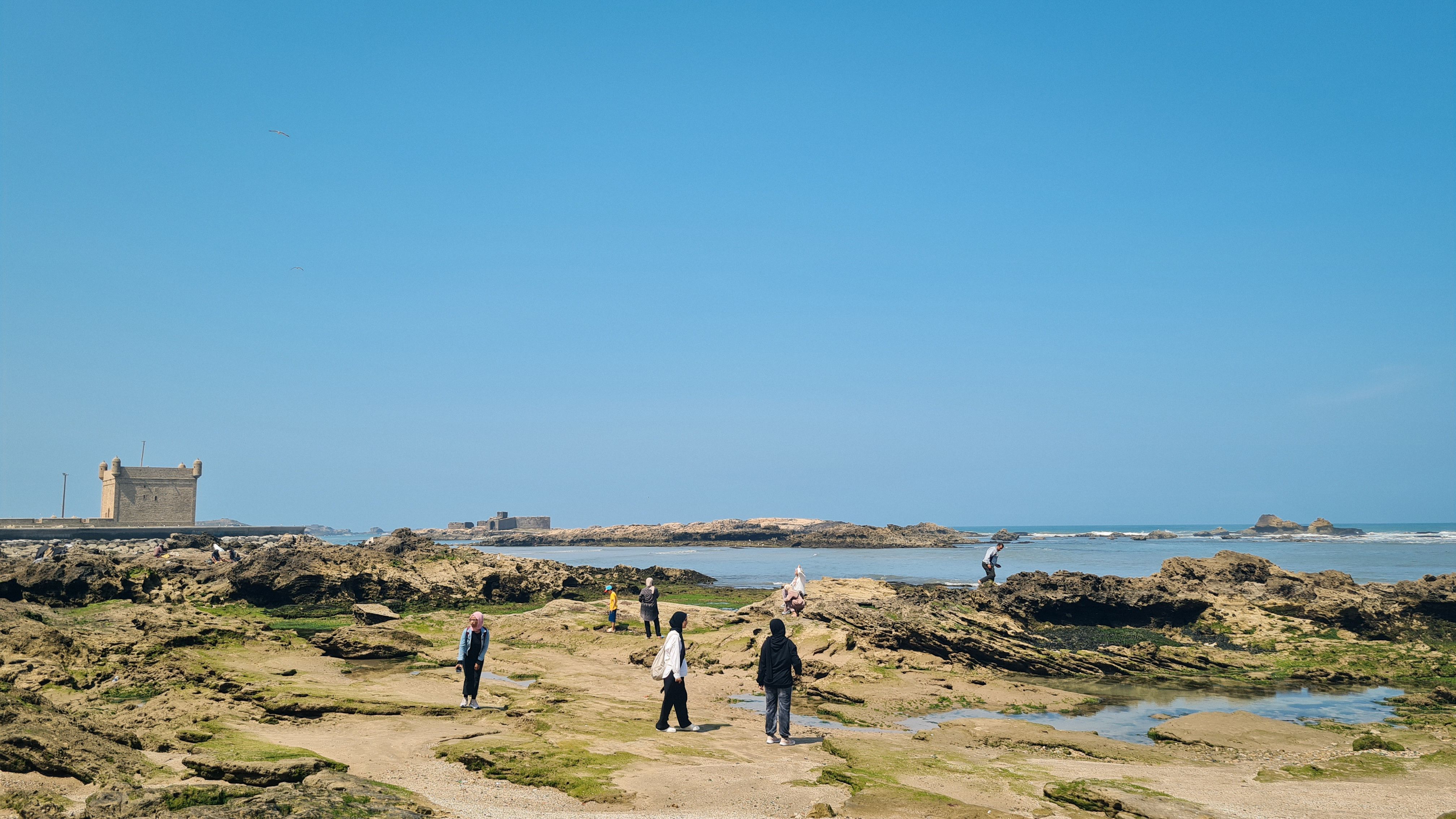 People walking over rocks and tide pools at low tide, with a stone bastion on the left, Essaouira