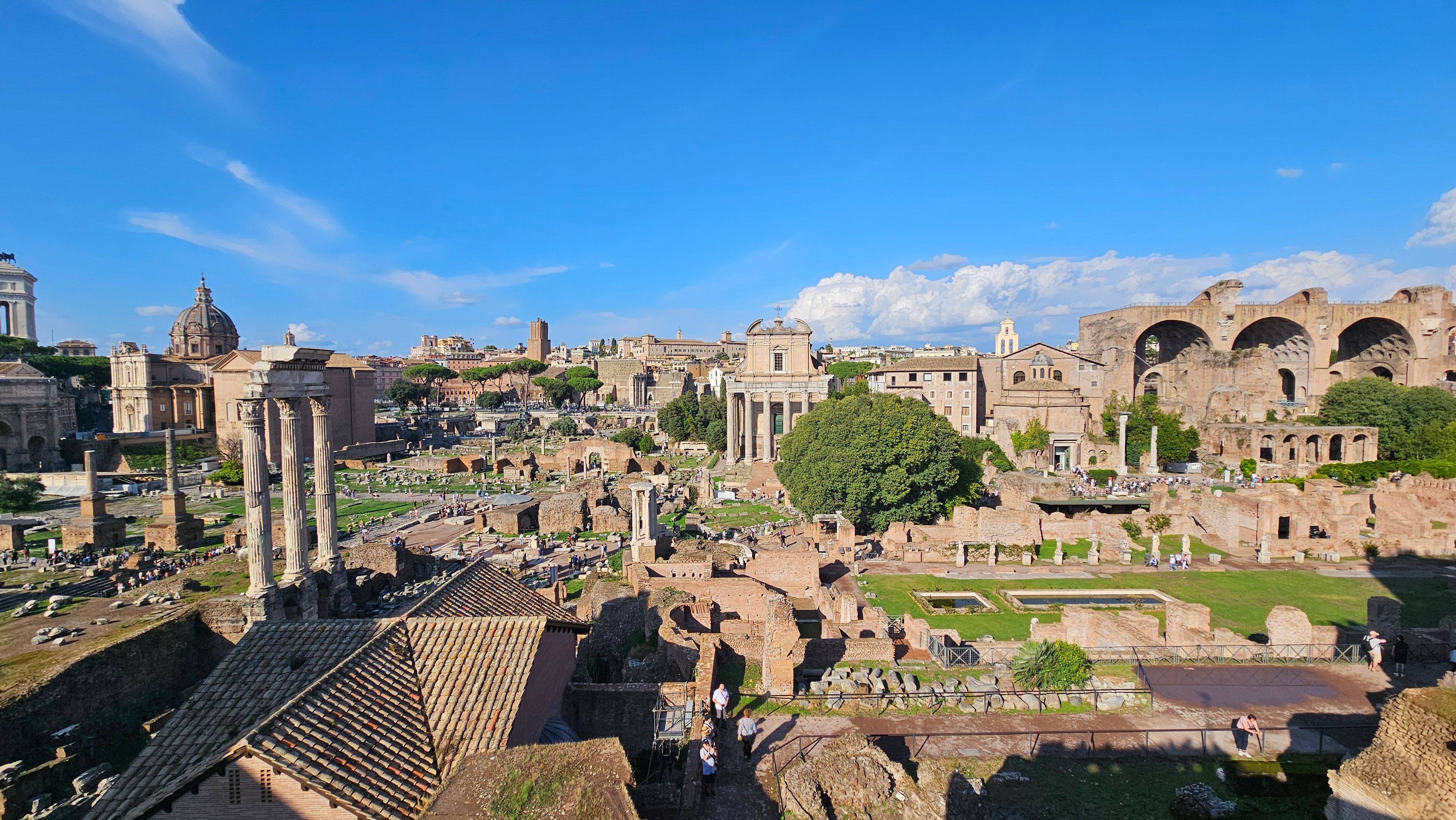 Wide view over the ruins of the Roman Forum under a blue sky, with three standing columns on the left, Rome