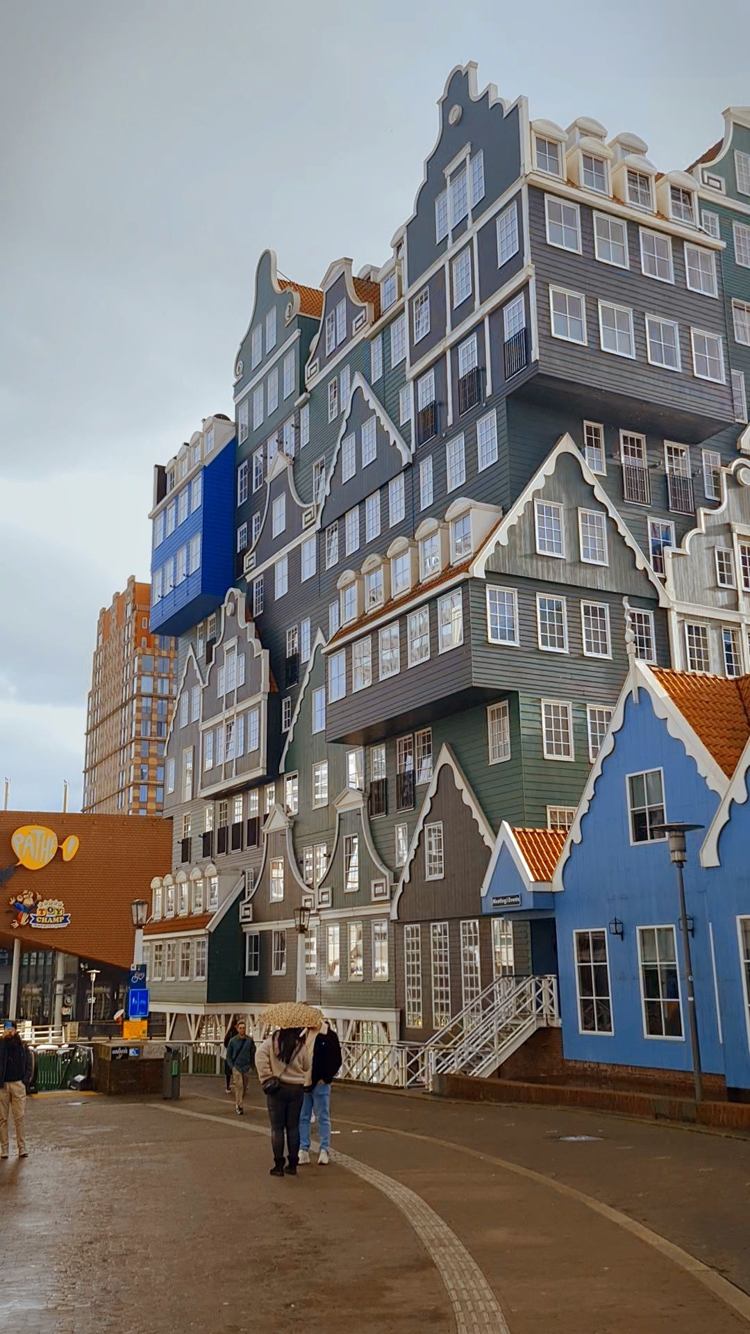 A hotel built from stacked green, grey and blue wooden gabled facades under an overcast sky, Zaandam