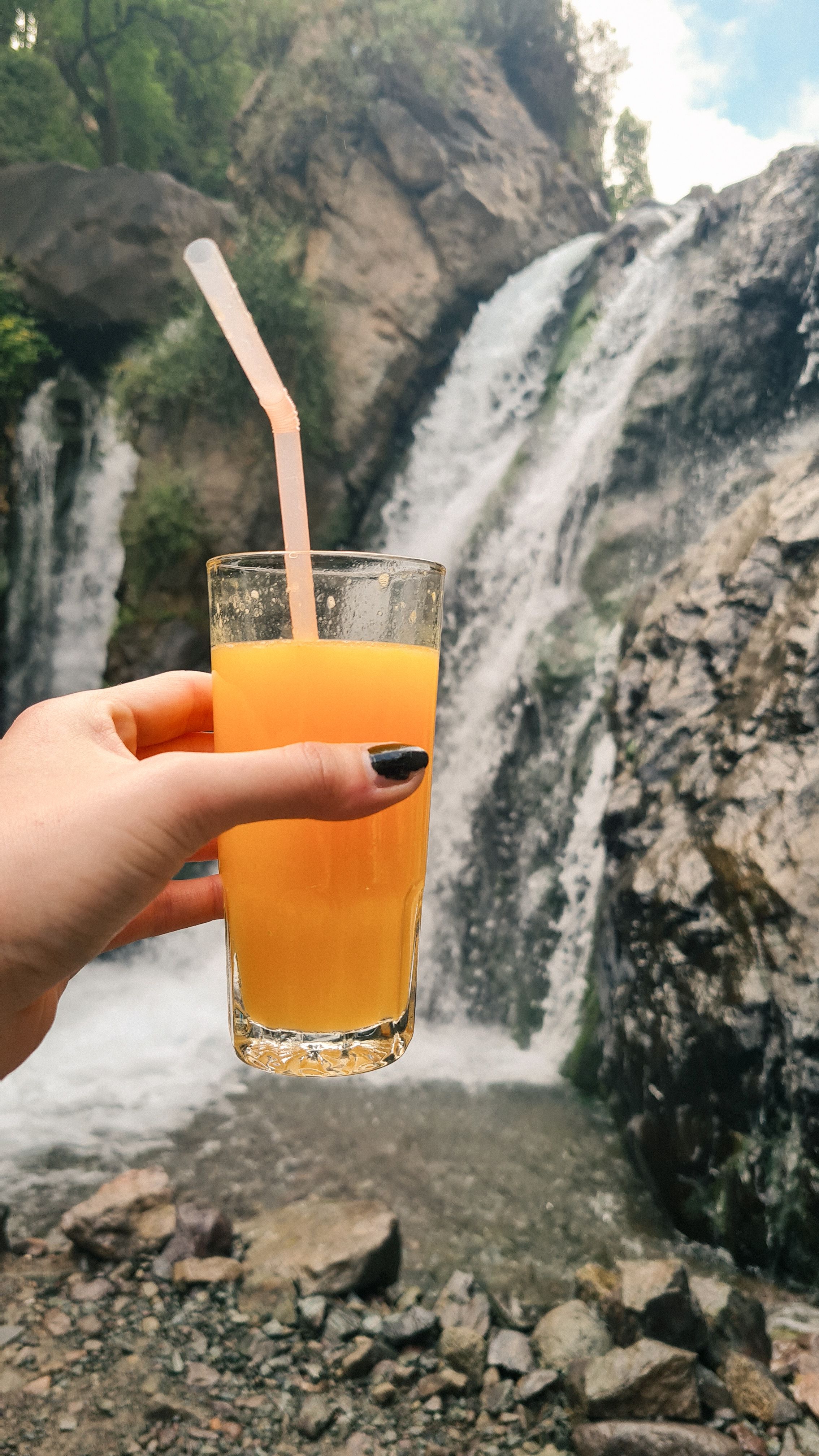 A hand holding a glass of orange juice in front of a waterfall in the Atlas Mountains, Imlil