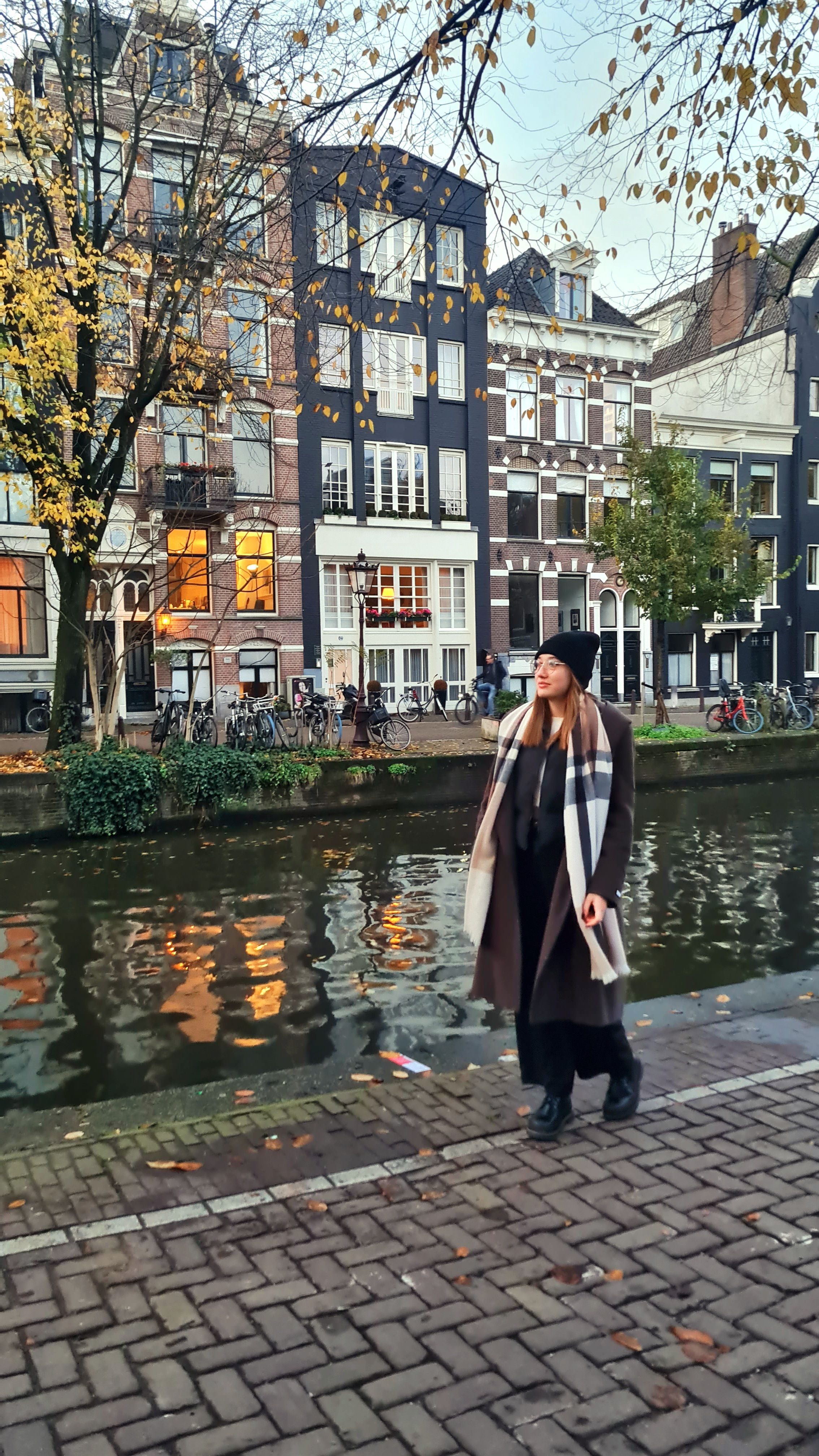 A woman in a long brown coat and black beanie walking along a canal lined with tall brick houses and yellow autumn trees, Amsterdam